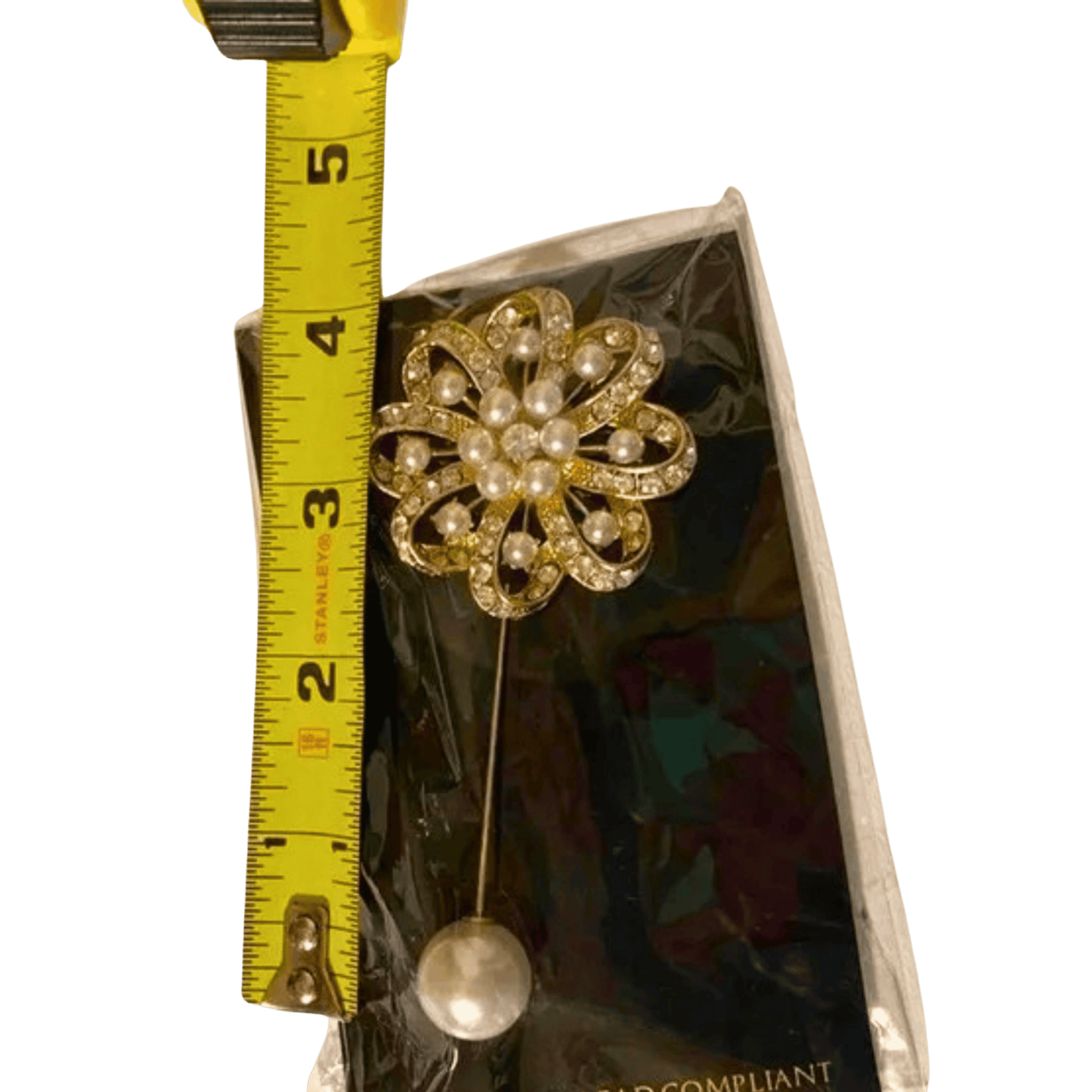 Gold brooch with pearl-like stones on a clear background, measured by a yellow tape measure.